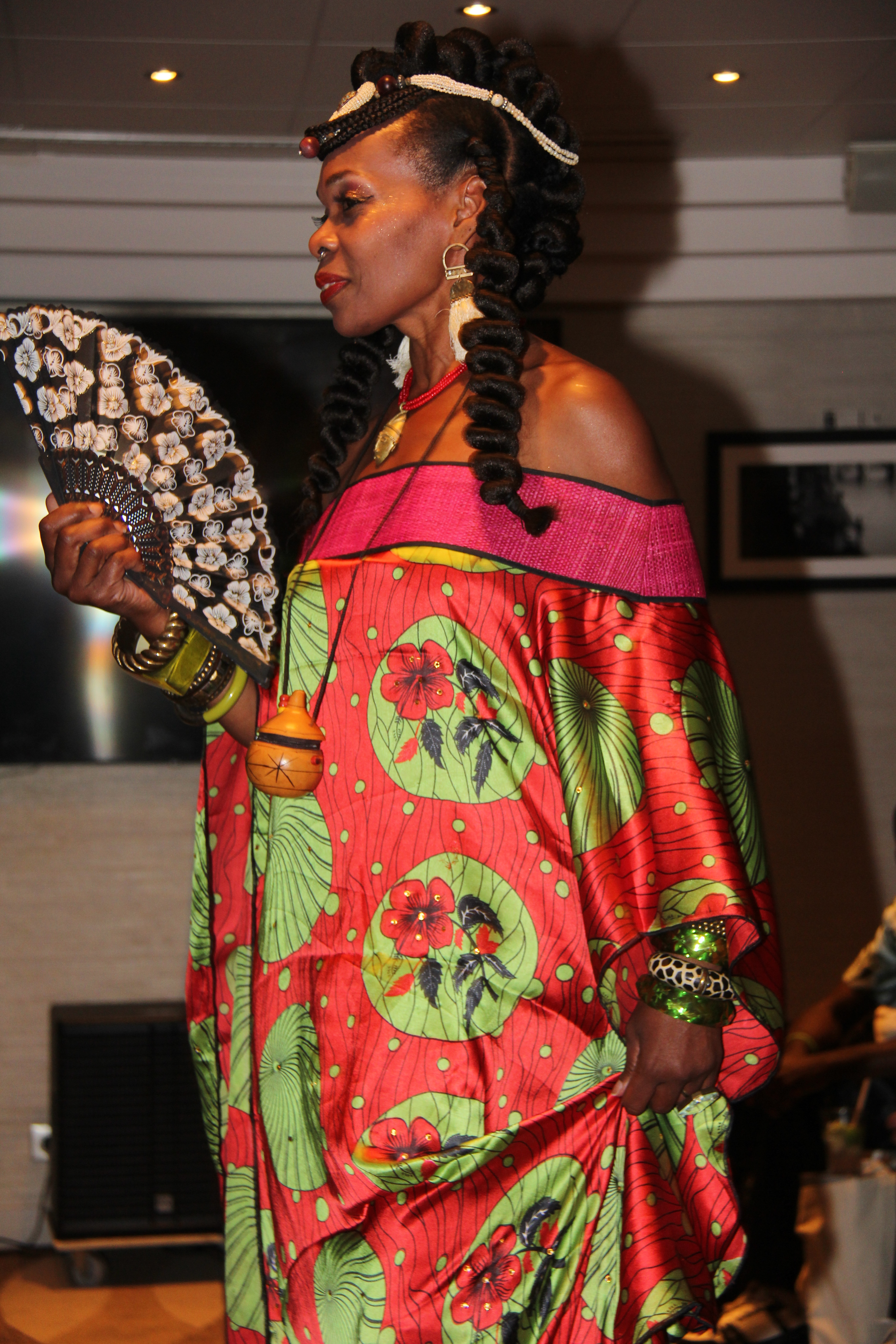 Femme portant une robe colorée à motifs floraux, tenant un éventail, avec des cheveux enroulés et des accessoires traditionnels.